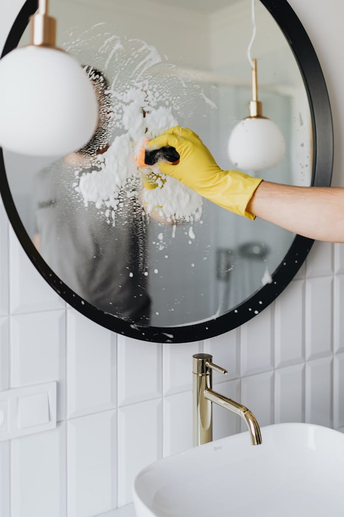 A person wearing a yellow glove cleans a bathroom mirror with foam spray.