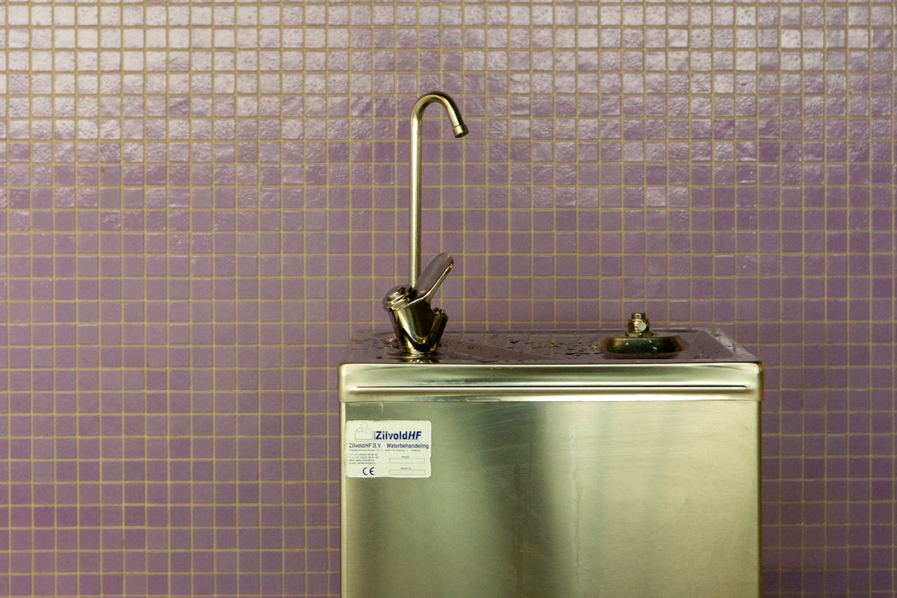 A stainless steel drinking fountain set against a purple mosaic tiled wall, urban environment.